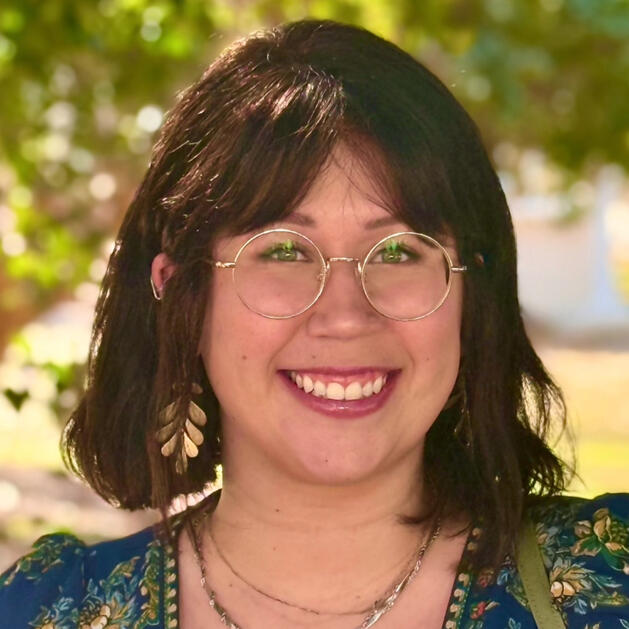 It's a headshot of a young woman smiling at the camera. She has dark brown shoulder length hair and bangs. She's wearing gold circular glasses and gold leaf earrings.