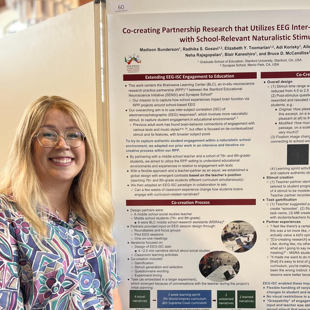It's a young blonde woman with glasses smiling at the camera. She's standing next to a scientific research poster.